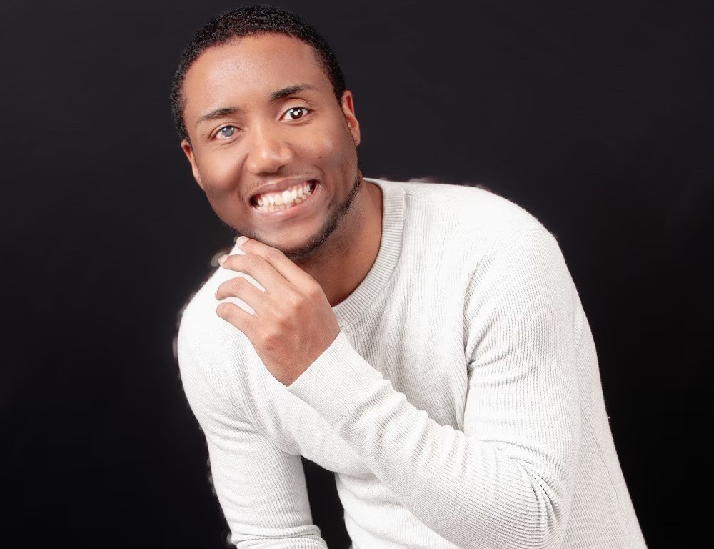 Cazerny Bussey smiling and posing in a light gray sweater against a black background, with his hand raised near his chin.