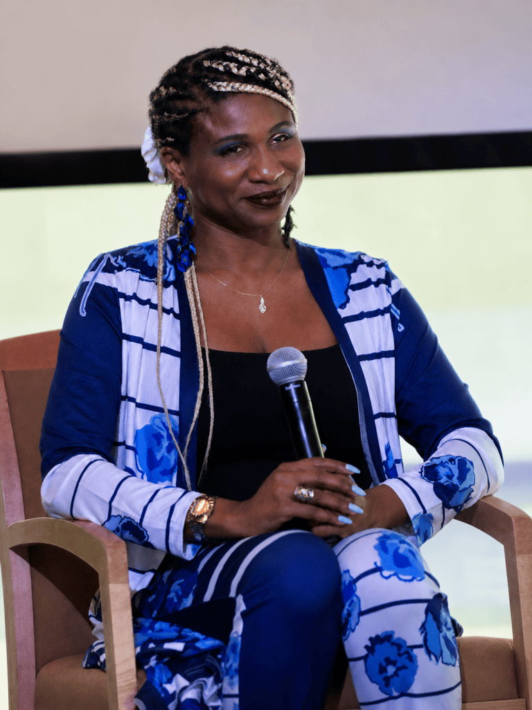 Lachi sits onstage during the Access in Focus panel wearing a blue shirt, with braids in her hair, speaking into a handheld microphone.