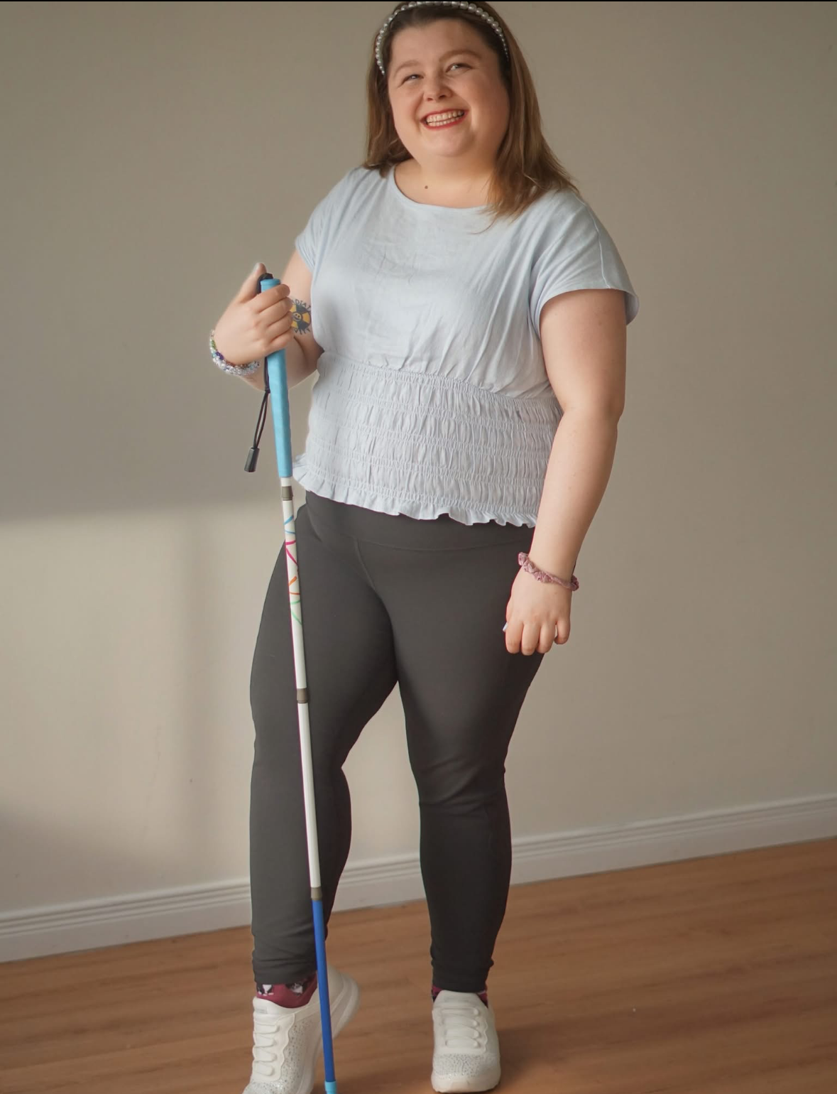 A smiling blind woman named Alicia Grace stands indoors holding a white mobility cane. She is wearing a light blue shirt, dark pants, white sneakers, and a jeweled headband. She is centered in the photograph and facing the camera.
