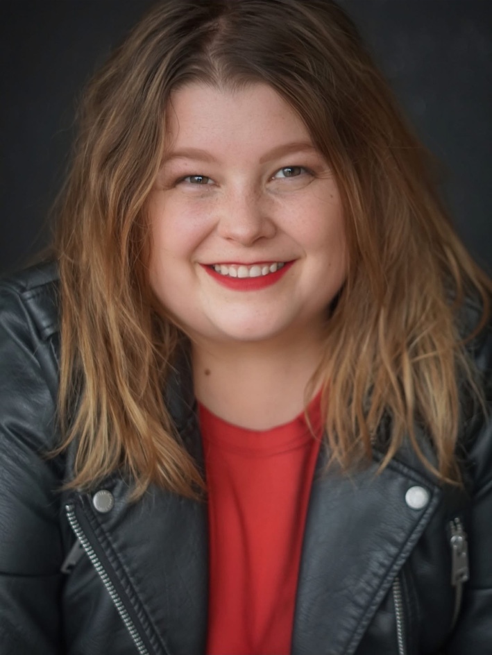 Alicia Grace smiles directly at the camera. Her long light-brown hair frames her face, and she is wearing a red top beneath a black leather jacket against a dark background.