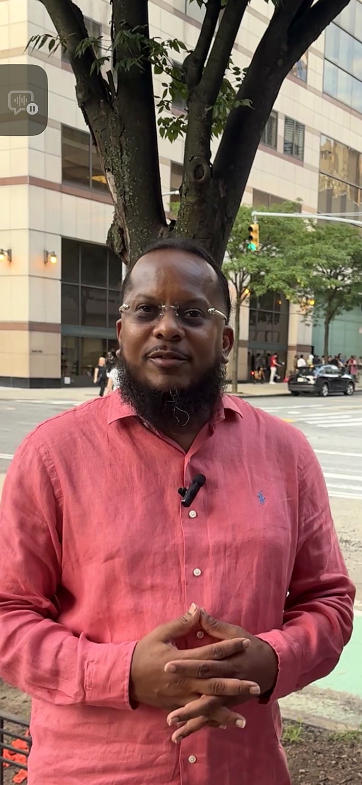 Hip-hop artist Special Ed stands outdoors on a city sidewalk in a bright coral-pink button-down shirt and glasses, facing the camera with his hands clasped. Buildings, trees, pedestrians and traffic appear softly out of focus behind him.