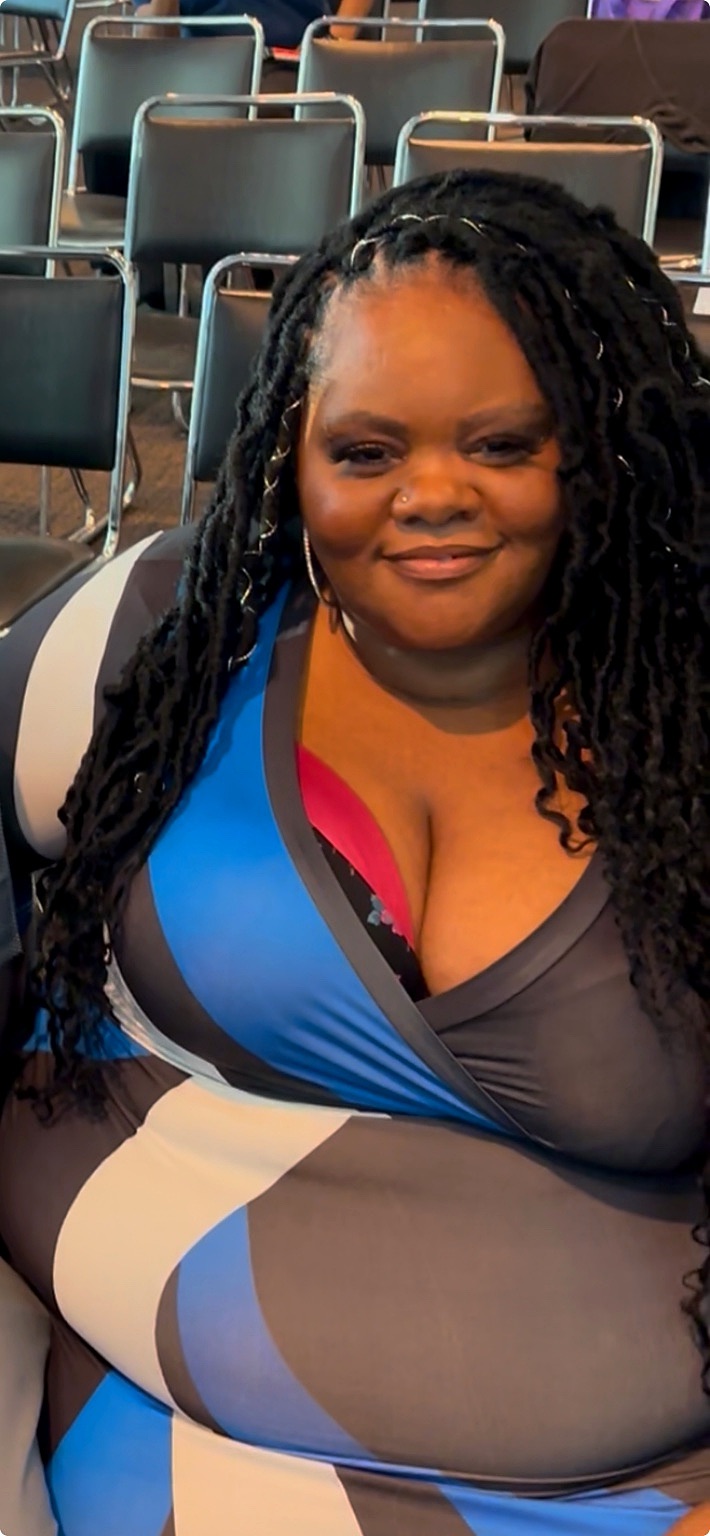 Nicole Harris sits smiling among rows of auditorium chairs at the Access in Focus event. She wears a patterned dark top and has long locs draped over her shoulders, with the event seating and other attendees visible around her.