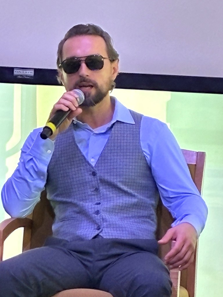 Petr Kucheryavyy sits indoors speaking into a handheld microphone during the Access in Focus panel. He wears glasses and a light-colored shirt, with another person seated beside him and the event space visible in the background.