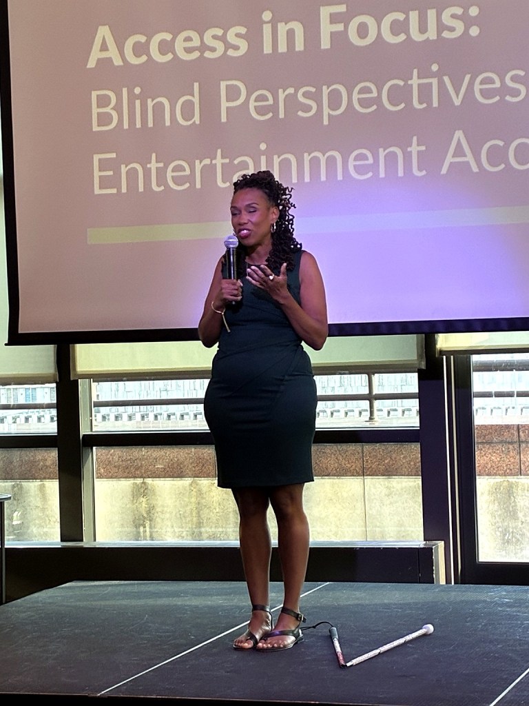 Kiana Glanton stands onstage speaking into a handheld microphone at Access in Focus. She wears a dark outfit in front of the event presentation screen, and a white cane rests on the stage near her feet.